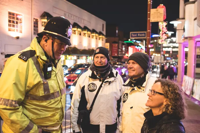 Two City Pastors volunteers in white jackets talking with a police officer in hi-vis and a colleague on a Birmingham street at night